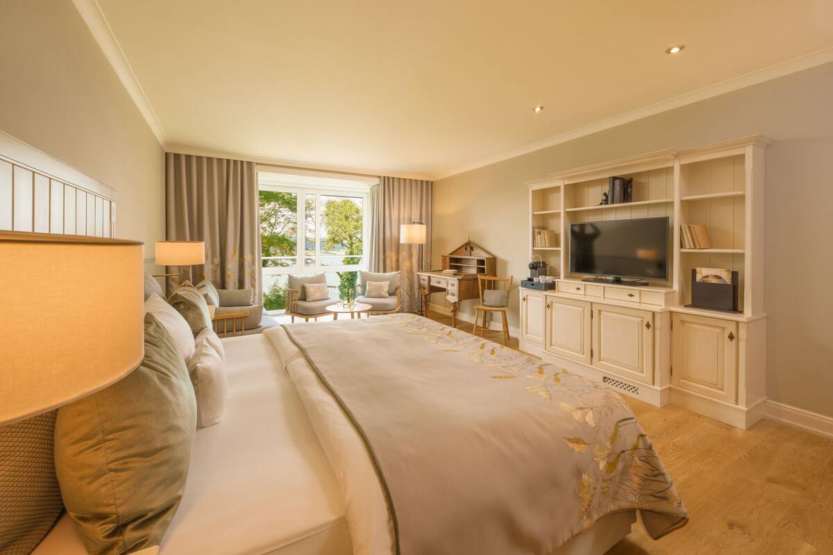 A hotel room with a large bed, a TV, a desk with a lamp, and a view of the outdoors.