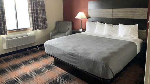 A hotel room with a large bed, a chair, a nightstand with a lamp, a window with curtains, and a wall-mounted air conditioner.