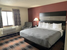 Quarto de hotel com uma cama grande, uma cadeira, mesa de cabeceira com abajur, janela com cortinas e ar condicionado fixado na parede.