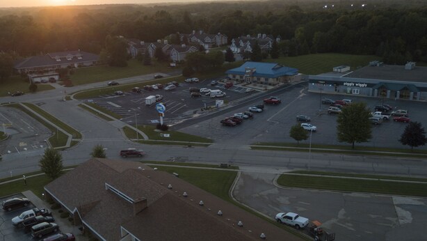 Une vue aérienne d’un parking rempli de voitures, d’un bâtiment avec un toit bleu et d’un grand champ en arrière-plan.