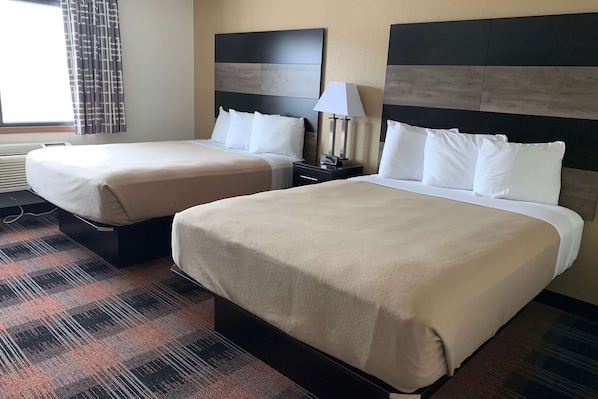 A hotel room with two beds, a nightstand with a lamp, a window with curtains, and a patterned carpet.