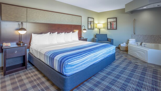 A hotel room with a large bed, a nightstand, a lamp, a chair, a bathroom with a bathtub, and a wall-mounted sink. - AmericInn by Wyndham Madison South (Madison)