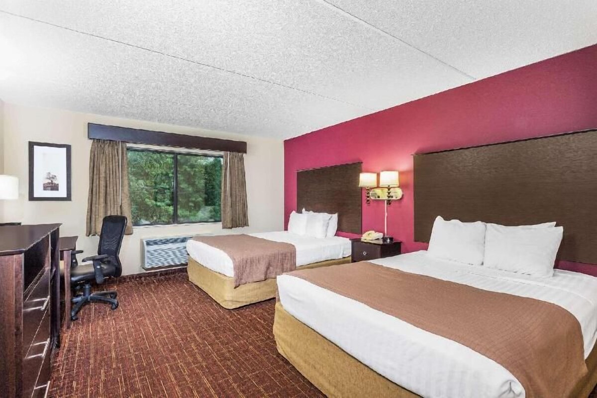 A hotel room with two beds, a desk, a chair, a window with curtains, and a lamp.
