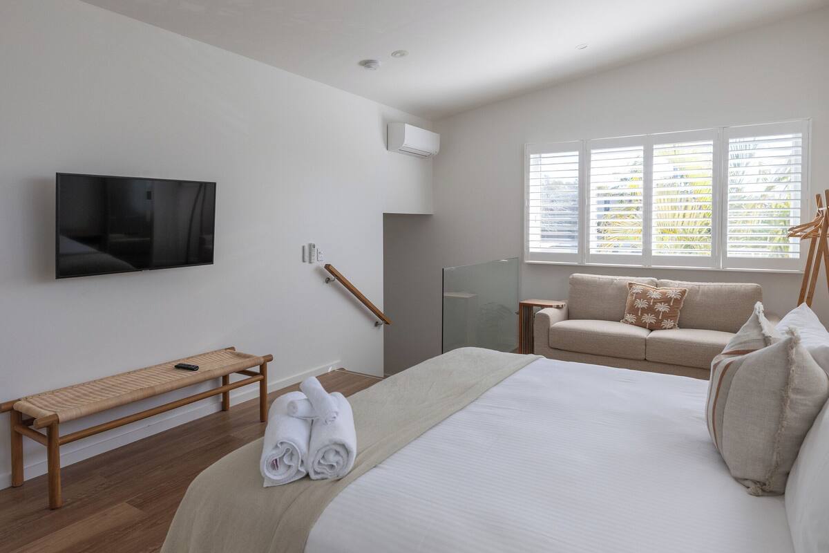 Quarto moderno com uma cama grande, um banco, uma TV e uma janela com persianas.