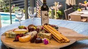 Une planche en bois garnie de différents fromages, de raisins et de crackers, accompagnée de deux verres à vin et d’une bouteille de vin, le tout disposé sur une table extérieure.