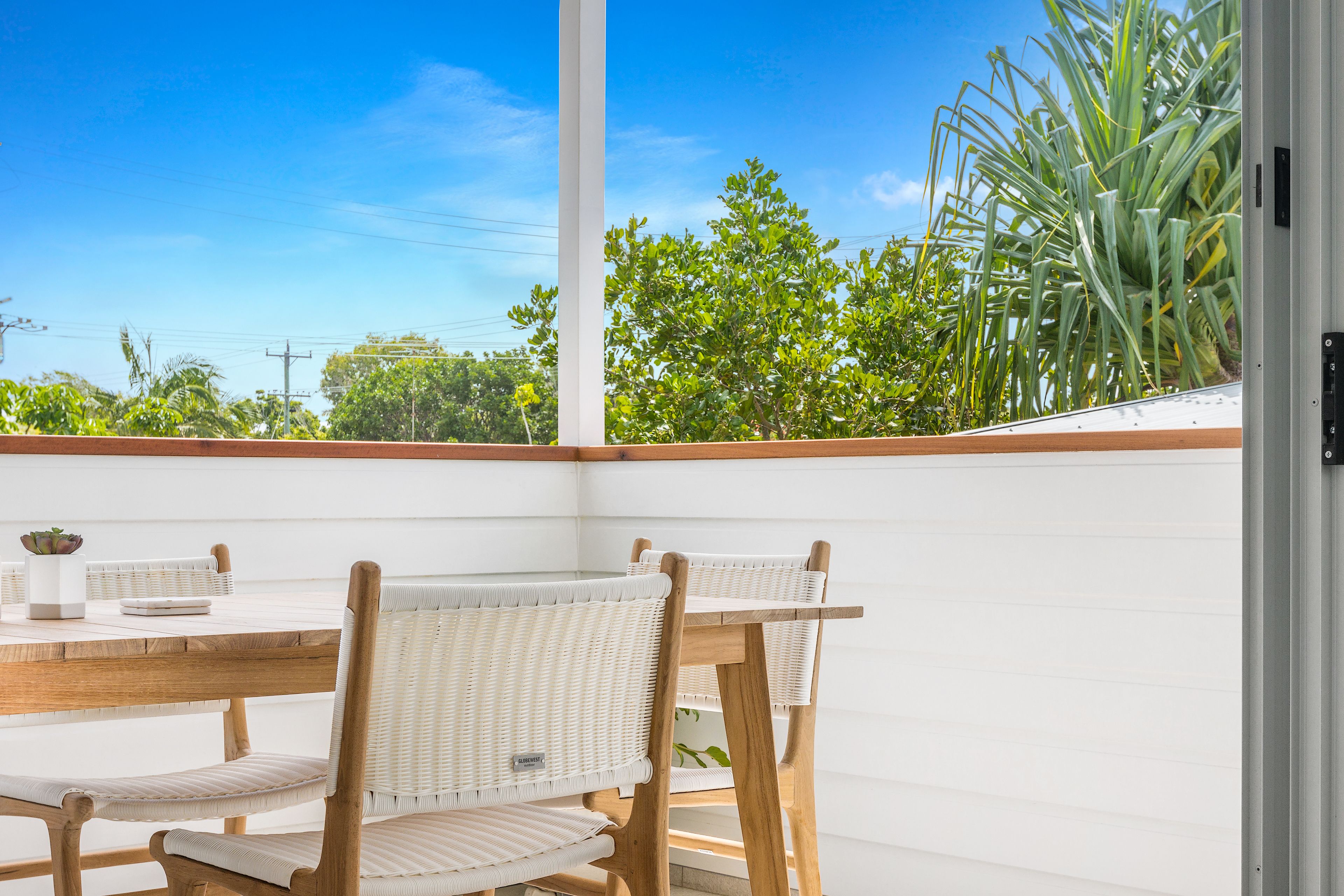 Un patio con mesa de madera y sillas de mimbre, con vistas a un jardín con diversas plantas y árboles.
