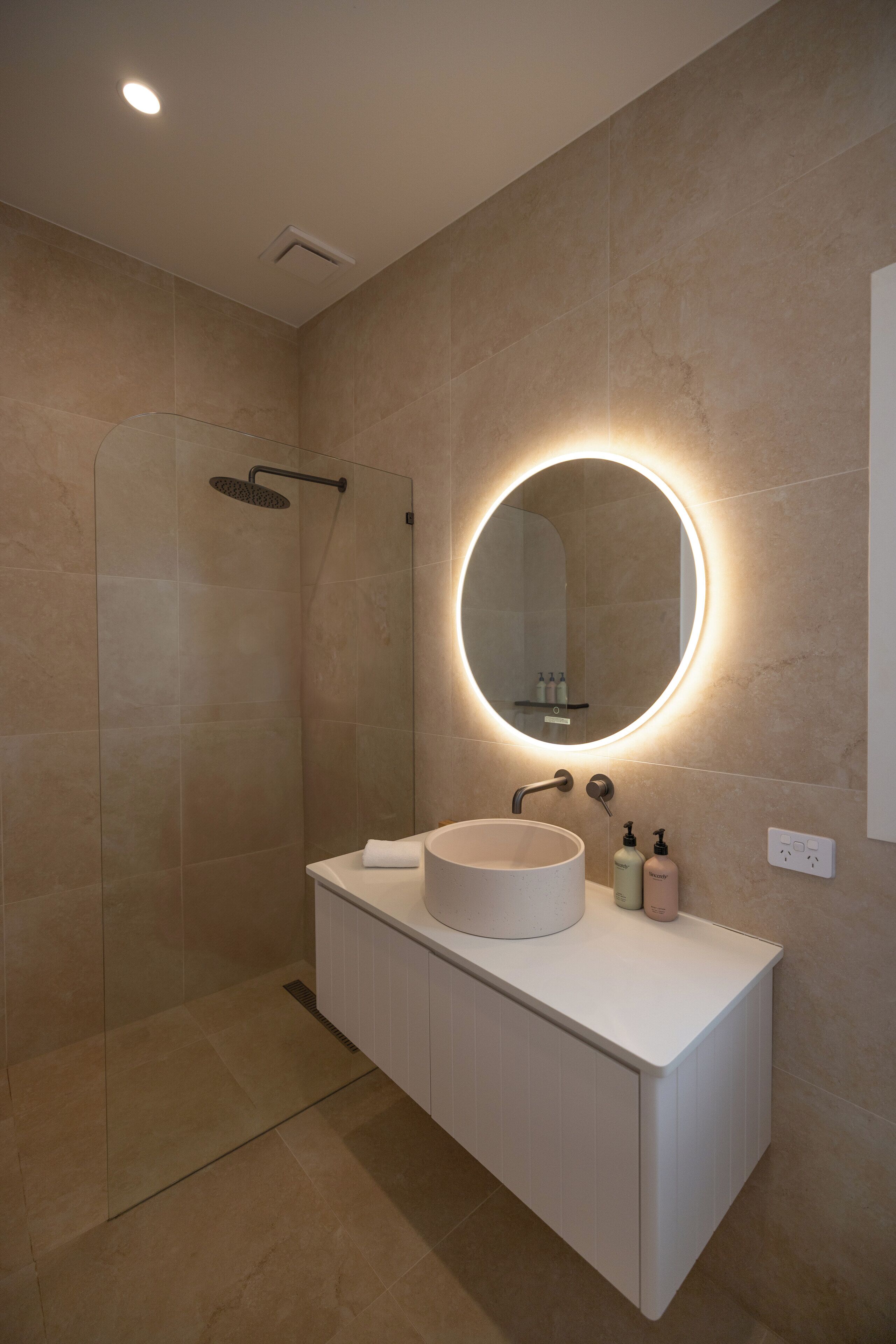 Une salle de bain moderne avec une douche à parois de verre, un grand miroir rond doté d’un éclairage indirect, un meuble-lavabo blanc et des toilettes.