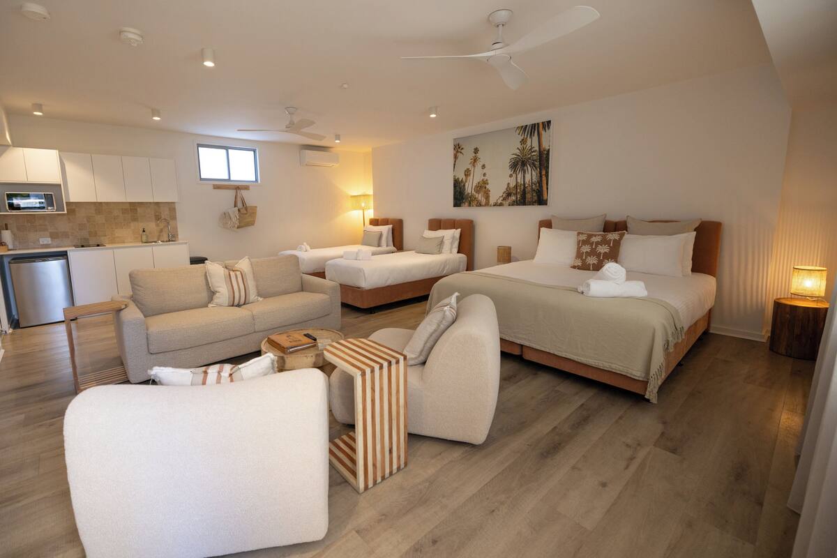 Quarto de hotel moderno com cozinha compacta, duas camas de solteiro, um sofá e um espaço de refeições.