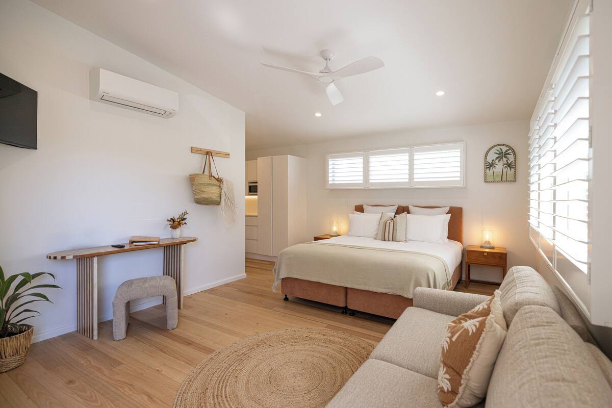 Quarto moderno com cama, mesas de cabeceira, escrivaninha, ventoinha no teto e janela com persianas.