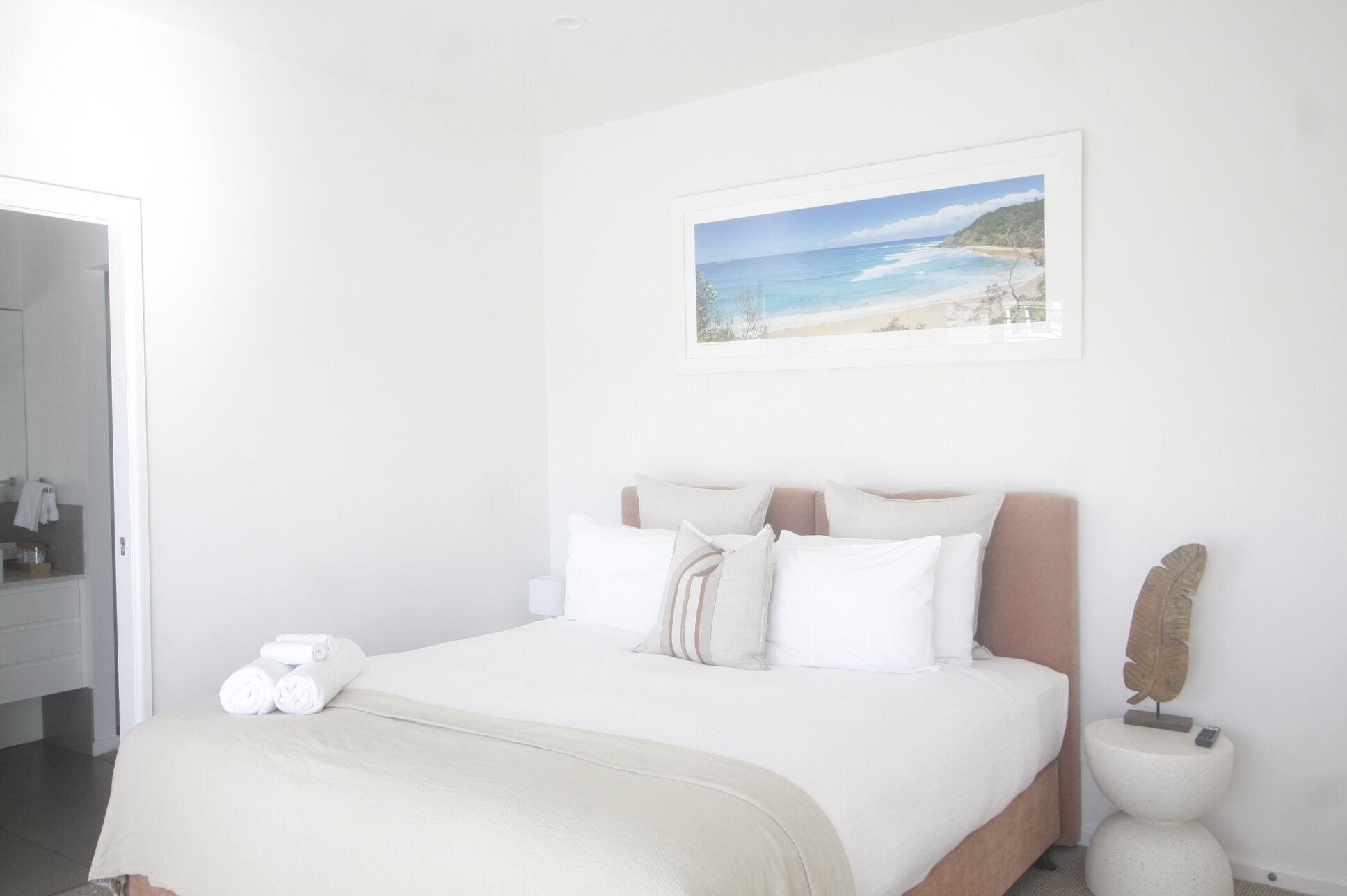 A bedroom with a bed, pillows, a painting of a beach, and a small decorative item on the nightstand.