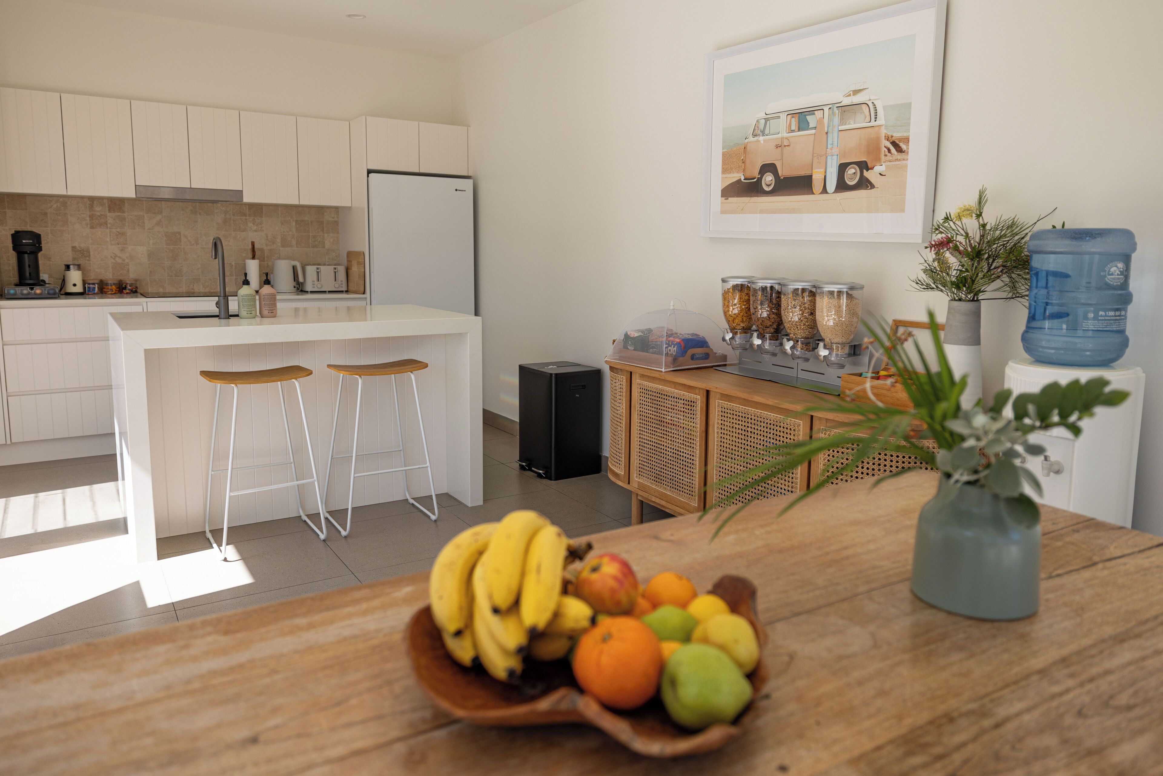 A modern kitchen with white cabinets, a central island with stools, a coffee maker, and a fruit bowl on the counter.