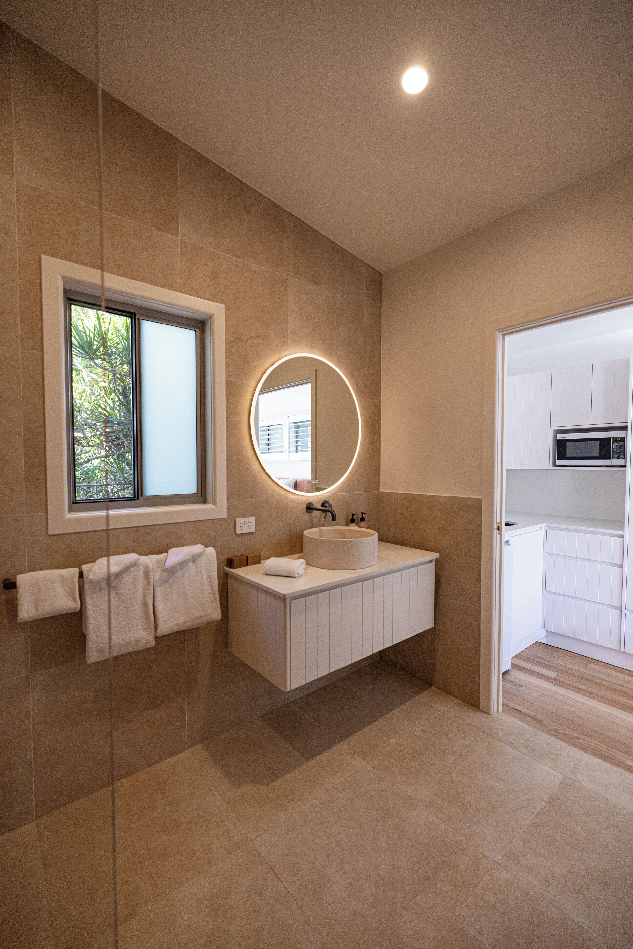 A modern bathroom with a large mirror, a round light above it, and a window with a view of greenery.
