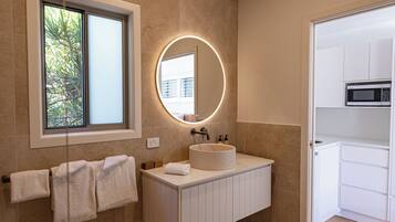 A modern bathroom with a large mirror, a round light above it, and a window with a view of greenery.