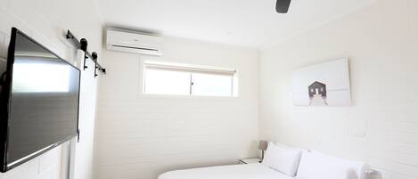 A hotel room with a bed, a TV mounted on the wall, a ceiling fan, an air conditioning unit, a window, and a nightstand with a lamp.