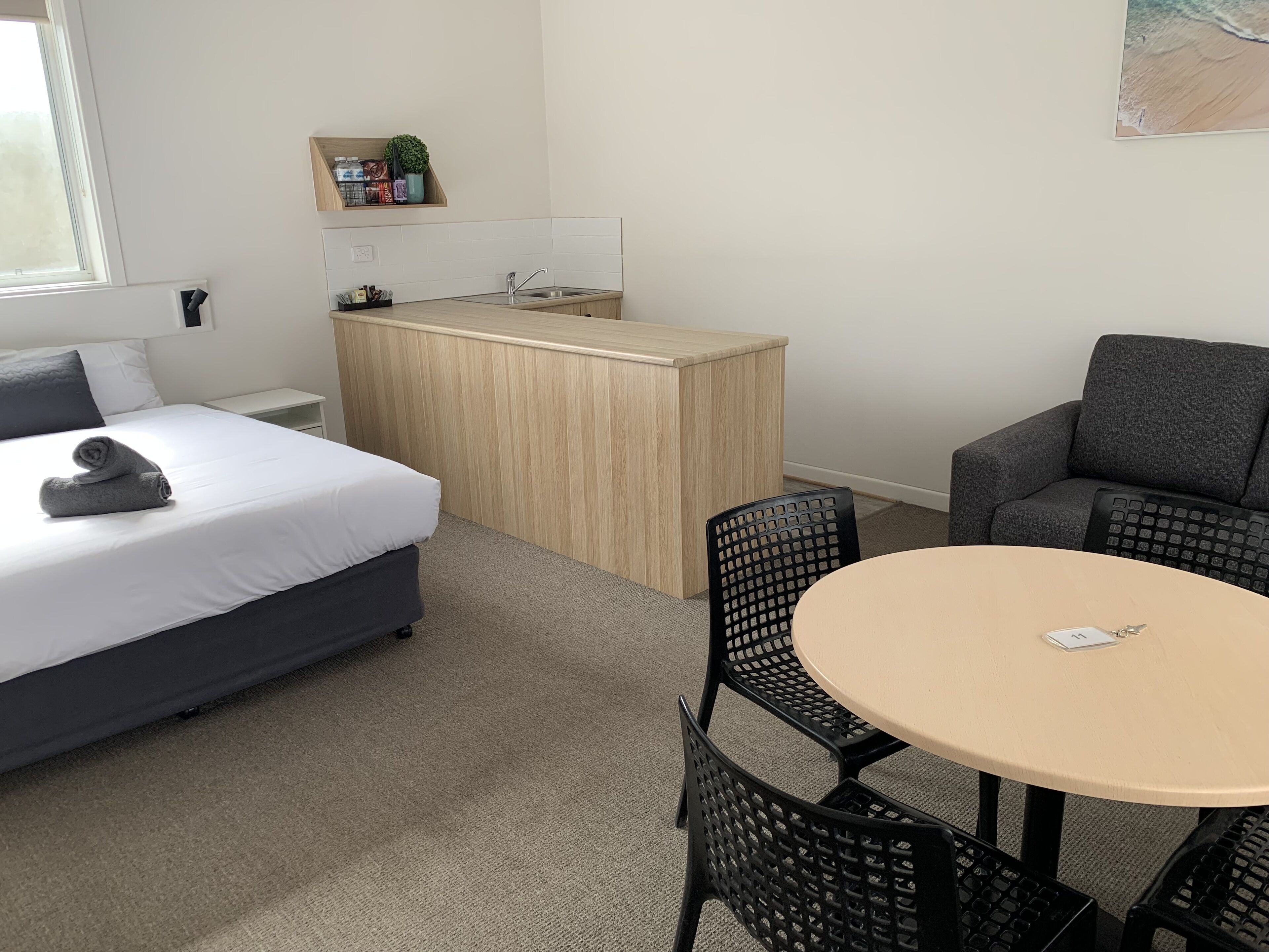 Habitación de hotel compacta con cama, kitchenette pequeña, mesa redonda con sillas y sofá gris.