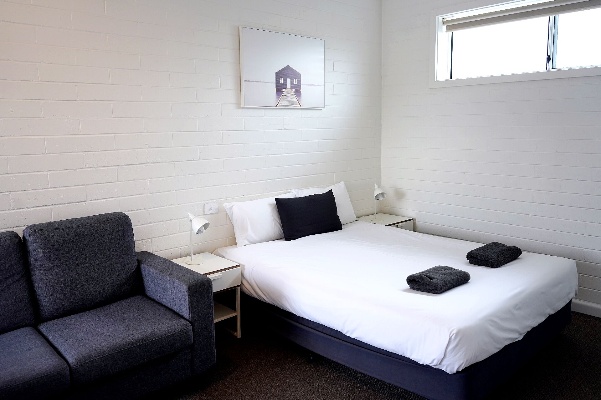 A hotel room with a bed, a chair, a nightstand, a lamp, and a window.