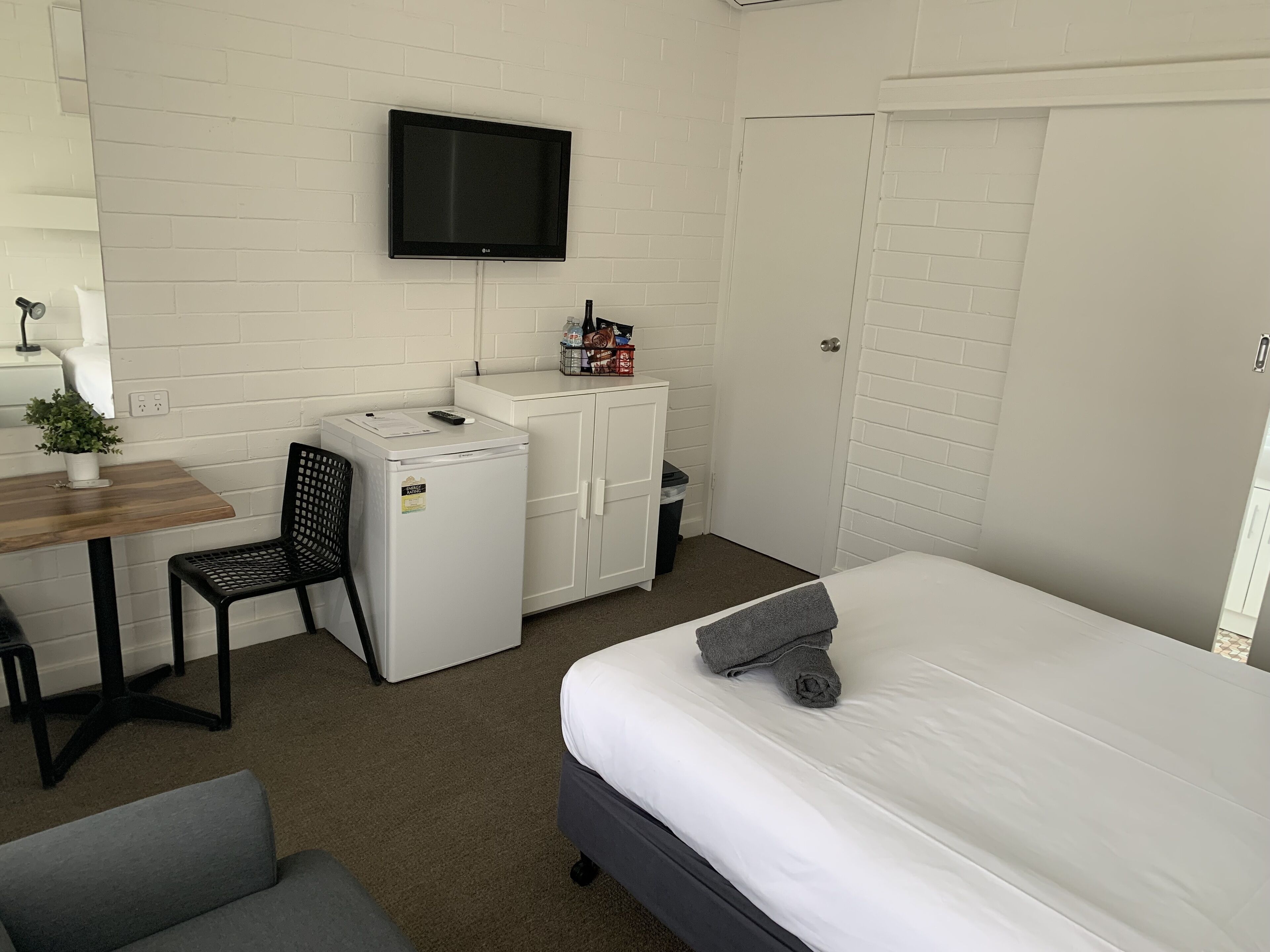 Habitación de hotel compacta con cama, escritorio y silla, un refrigerador pequeño, televisor montado en la pared y armario.