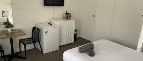 A compact hotel room with a bed, a desk with a chair, a small refrigerator, a TV mounted on the wall, and a wardrobe.