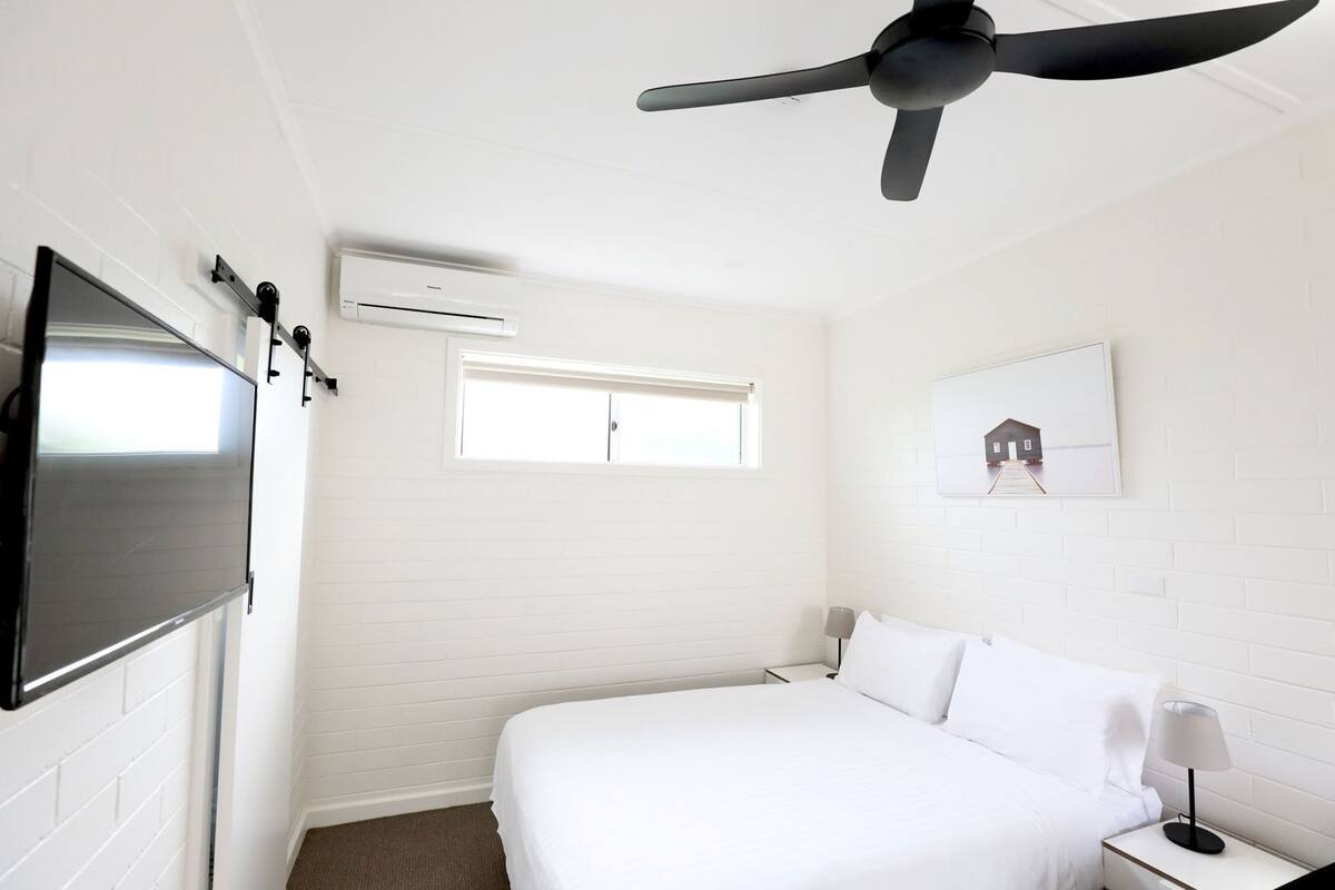 Habitación de hotel con cama, ventilador de techo, aire acondicionado, ventana y un cuadro en la pared.