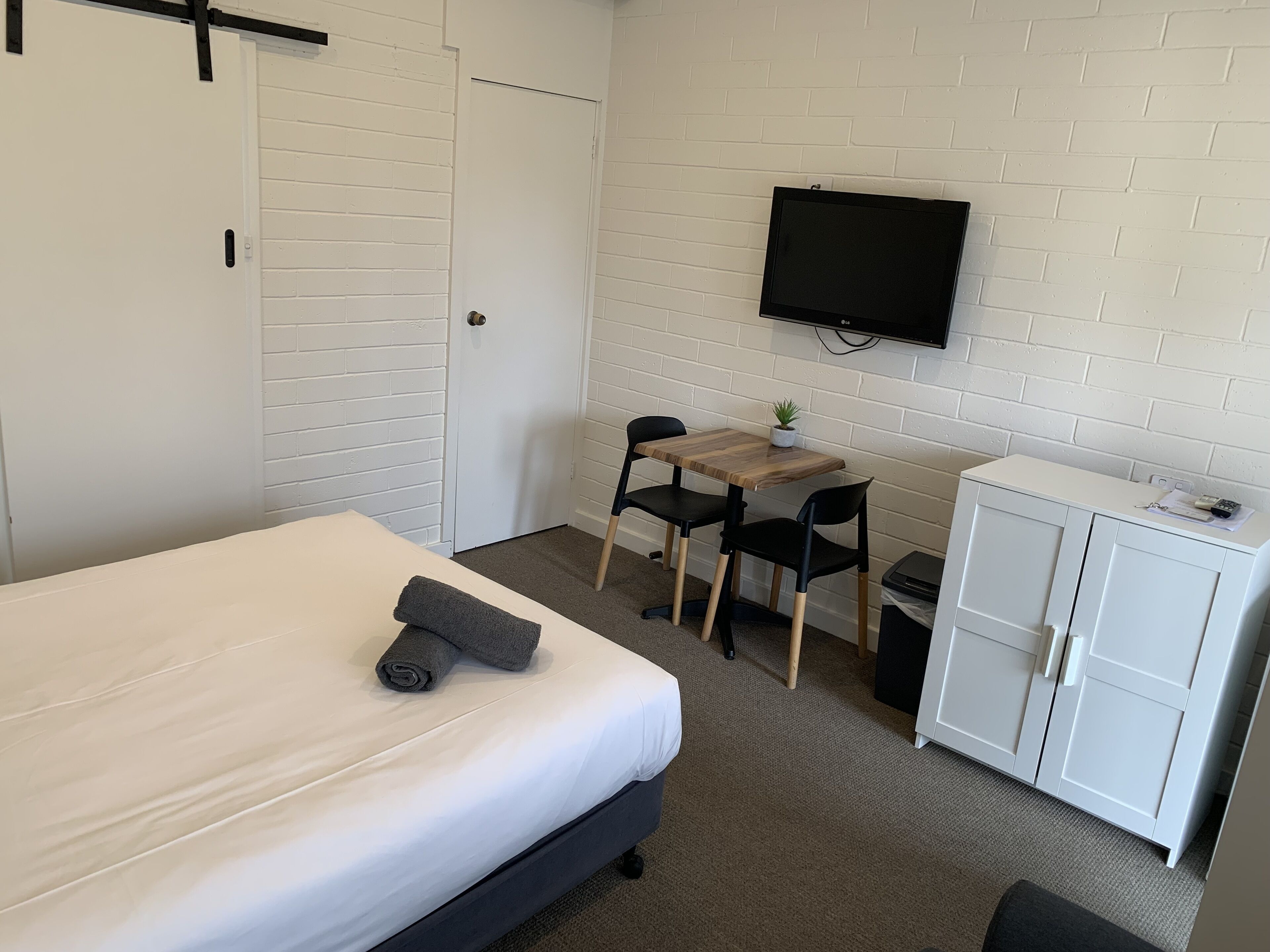 Una habitación de hotel con cama, escritorio con silla, televisor montado en la pared y armario.