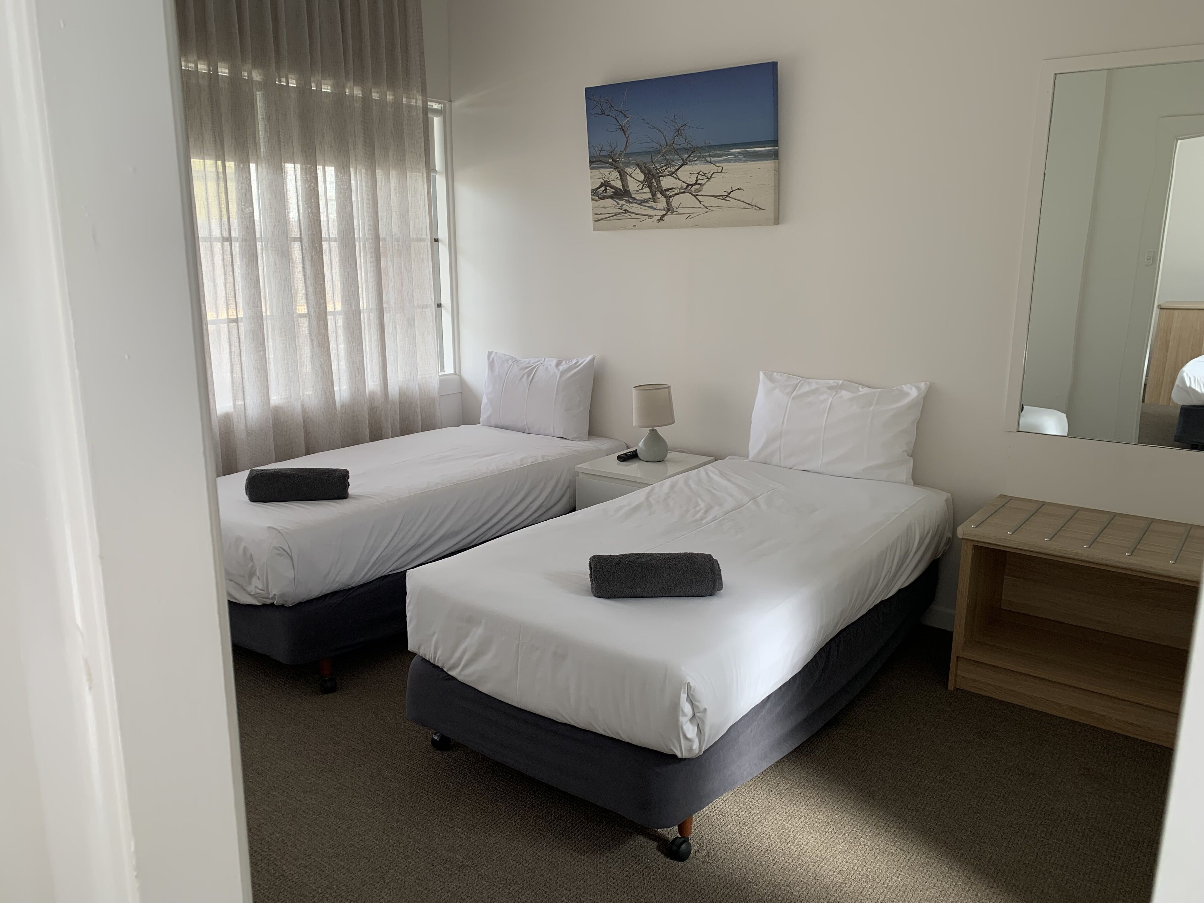 Habitación de hotel con dos camas individuales, una mesita de noche con lámpara, un pequeño estante de madera y un cuadro con una escena de playa.
