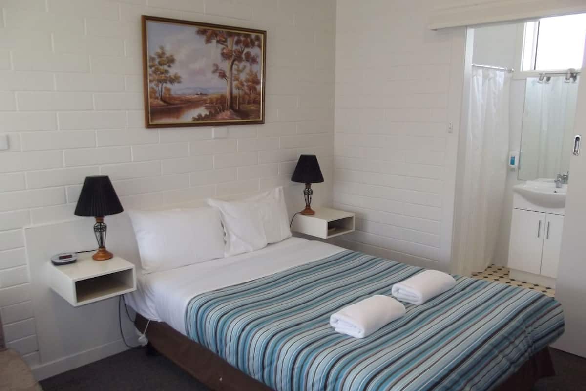 Habitación de hotel con cama, dos mesitas de noche con lámparas, un cuadro en la pared, un baño con lavamanos y espejo, y una ventana.