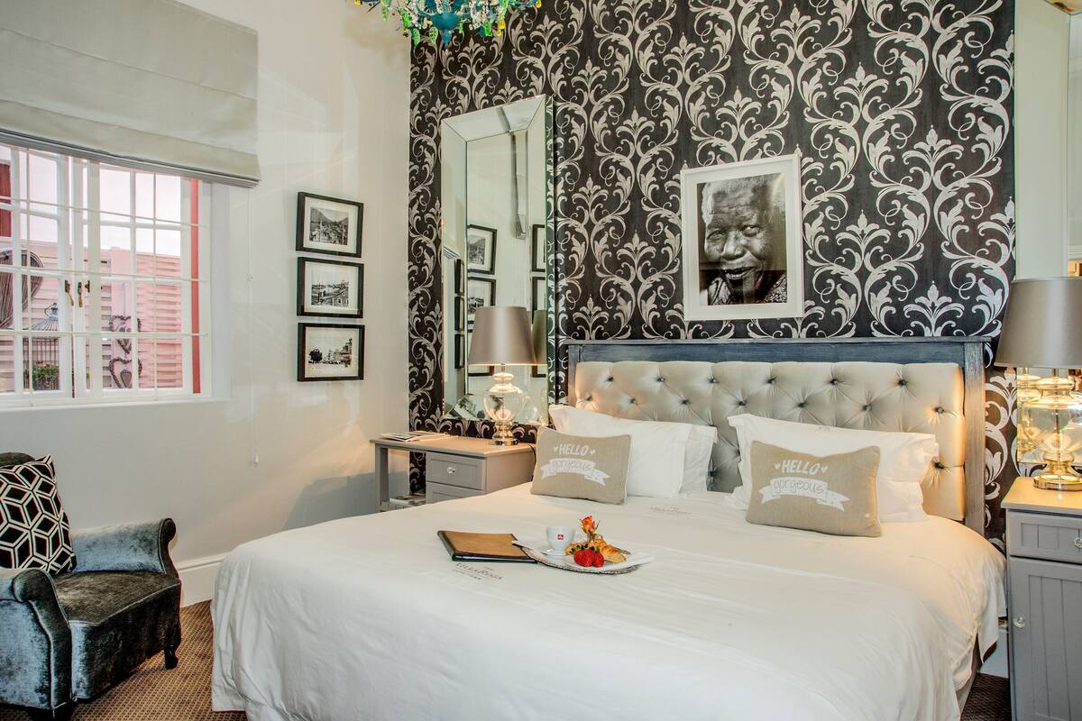 Un dormitorio ordenado con una cama grande, una mesita de noche con lámpara, una silla y una ventana con persianas.
