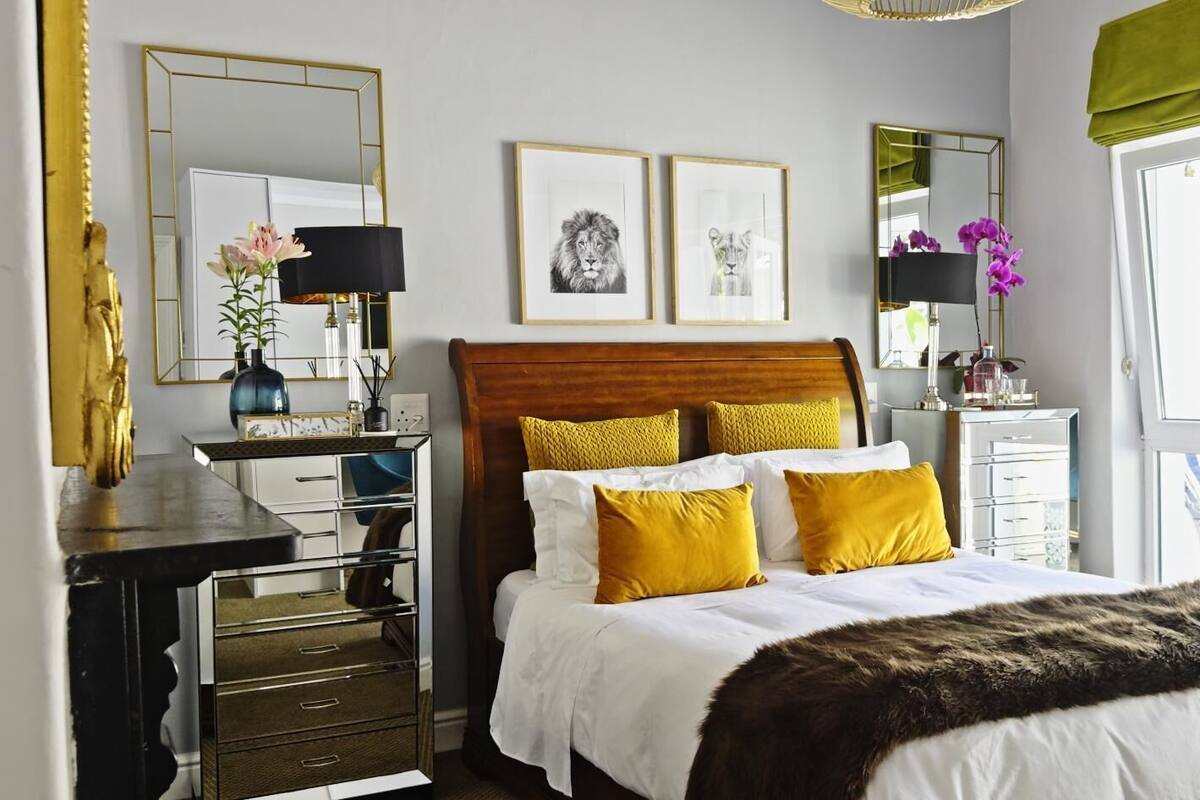 Un dormitorio con cabecera de madera, ropa de cama blanca y almohadas amarillas. Hay dos cuadros en la pared, un espejo y un escritorio con una lámpara.