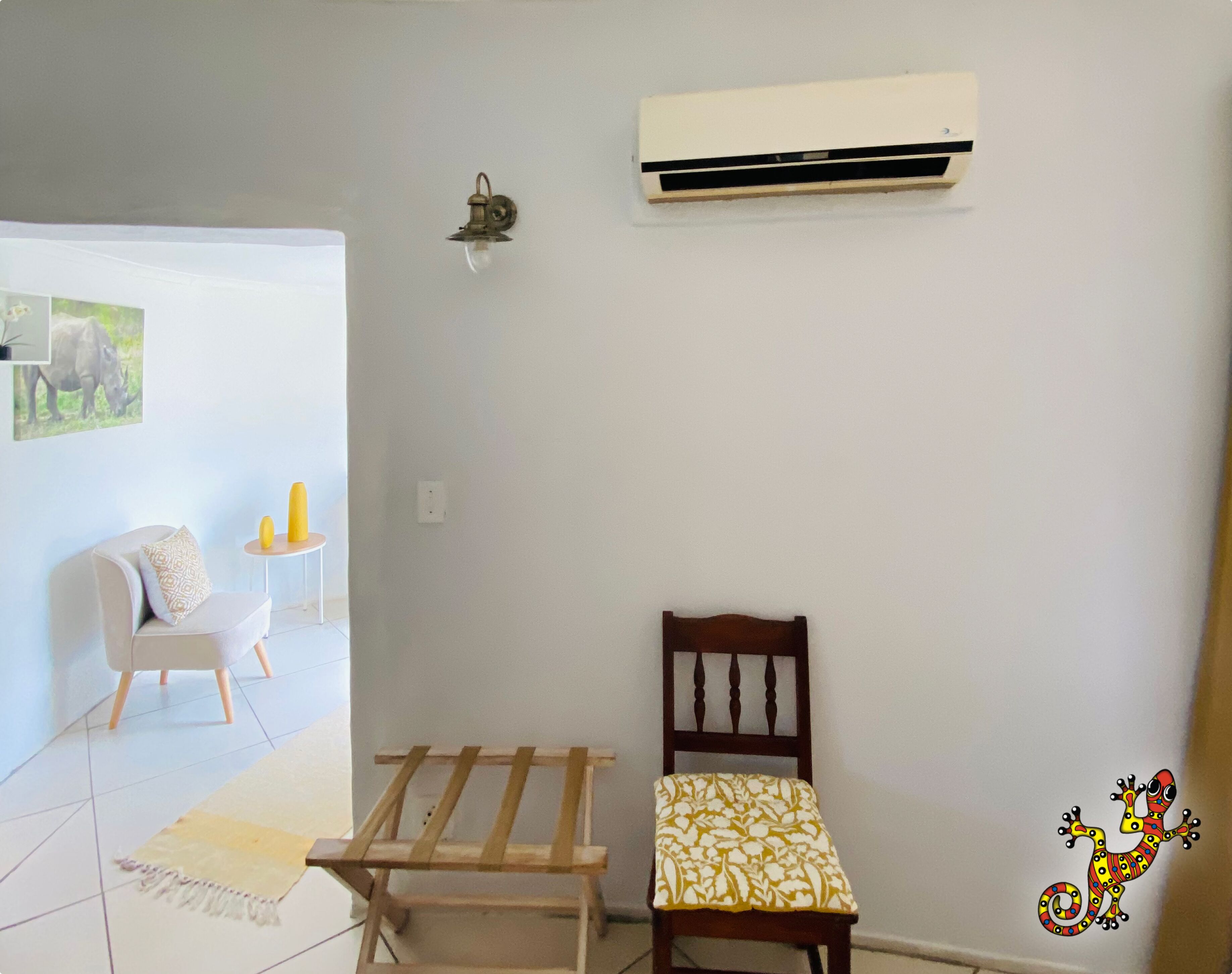 Una habitación con un marco de cama de madera, una pequeña silla de madera, un sofá blanco, una mesita redonda con una vela amarilla y un equipo de aire acondicionado en la pared.