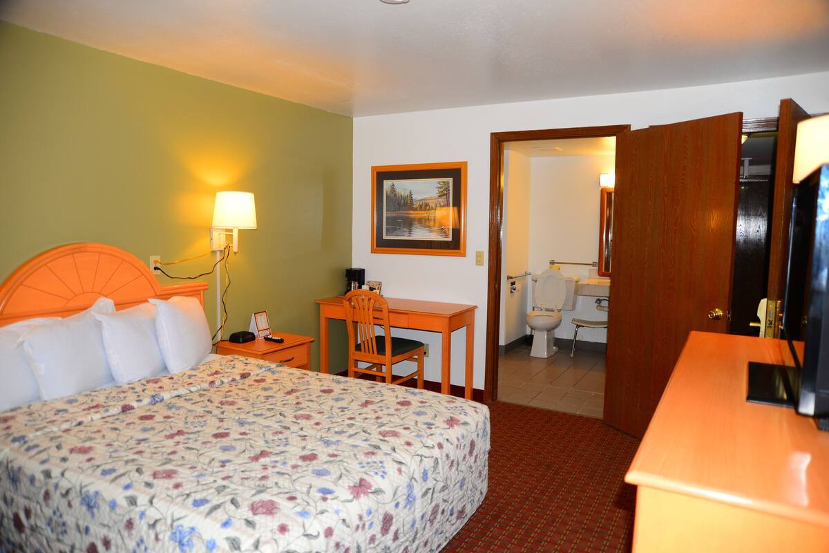 Habitación de hotel con una cama, un escritorio, una silla, una lámpara y un baño visible a través de una puerta abierta.