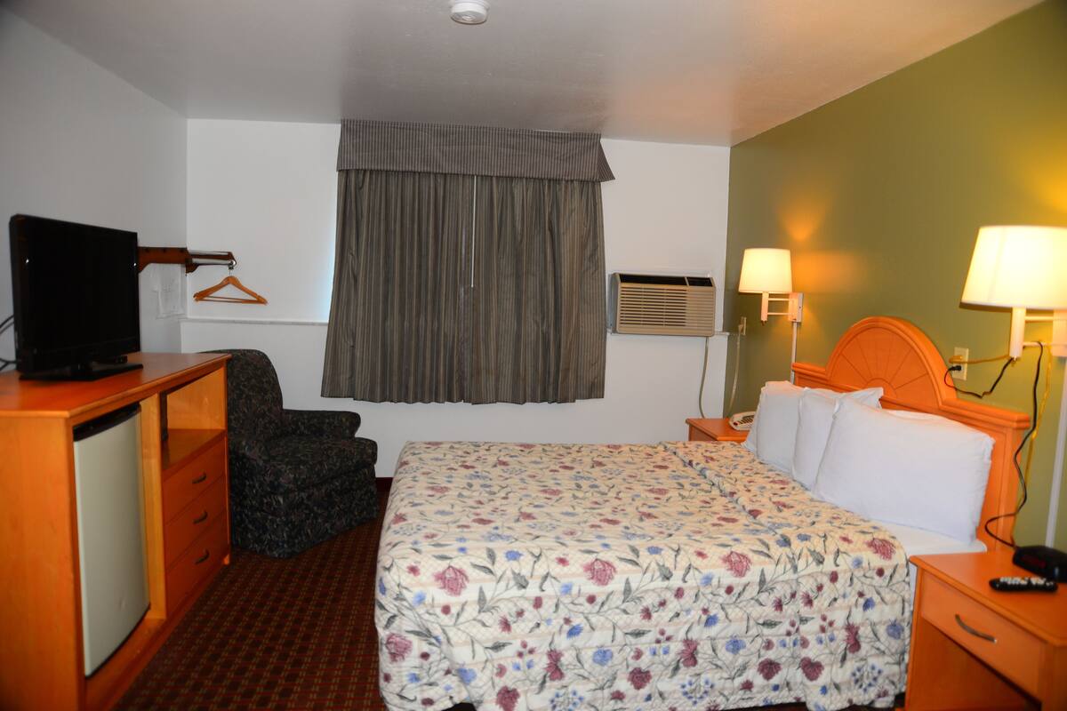 Habitación de hotel con cama, escritorio con televisor, una silla, armario, ventana con cortinas, unidad de aire acondicionado y lámparas de noche.