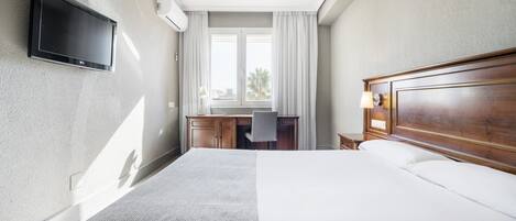 A hotel room with a bed, desk, chair, TV, and an air conditioning unit.