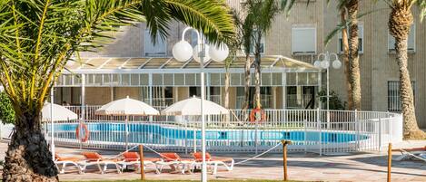 A swimming pool area with lounge chairs and umbrellas, surrounded by palm trees and a building with a modern design.