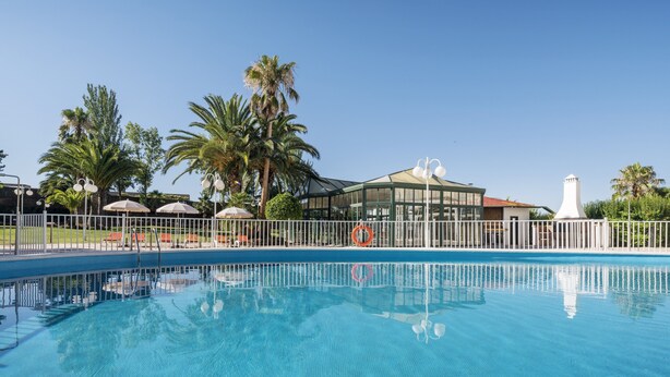 A clear blue pool with a white fence, palm trees, and a building with a glass facade. - Hotel ILUNION Las Lomas (Merida)