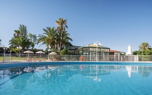 A clear blue pool with a white fence, palm trees, and a building with a glass facade. - Hotel ILUNION Las Lomas (Merida)