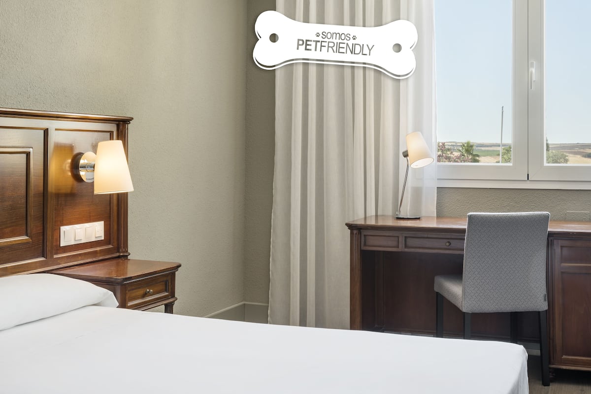 Ein Hotelzimmer mit Bett, Schreibtisch, Stuhl, Lampe und einem fenster mit Vorhängen.