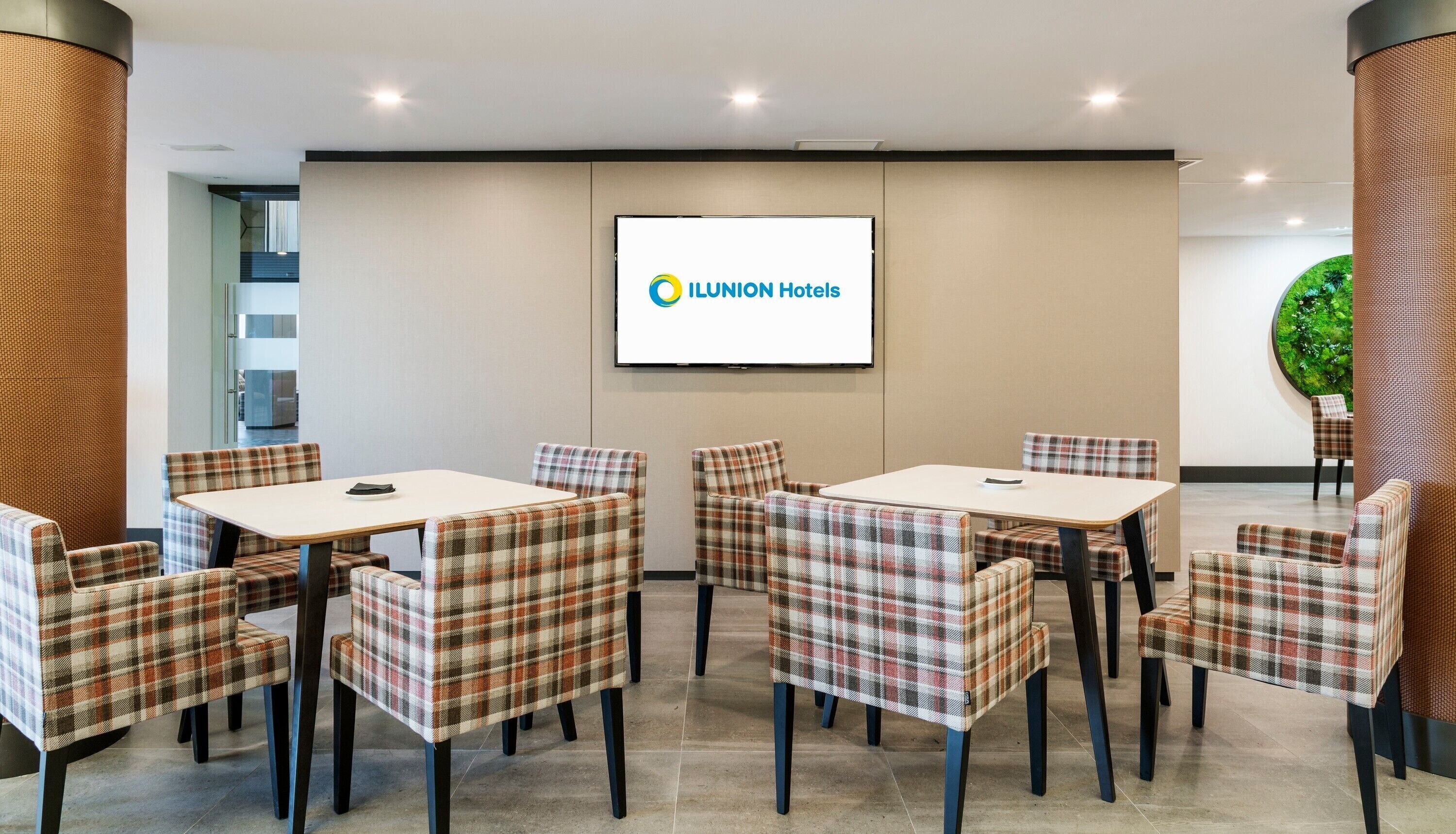 A conference room with plaid chairs and tables, a large screen displaying the ILUNION Hotels logo, and a green wall decoration.