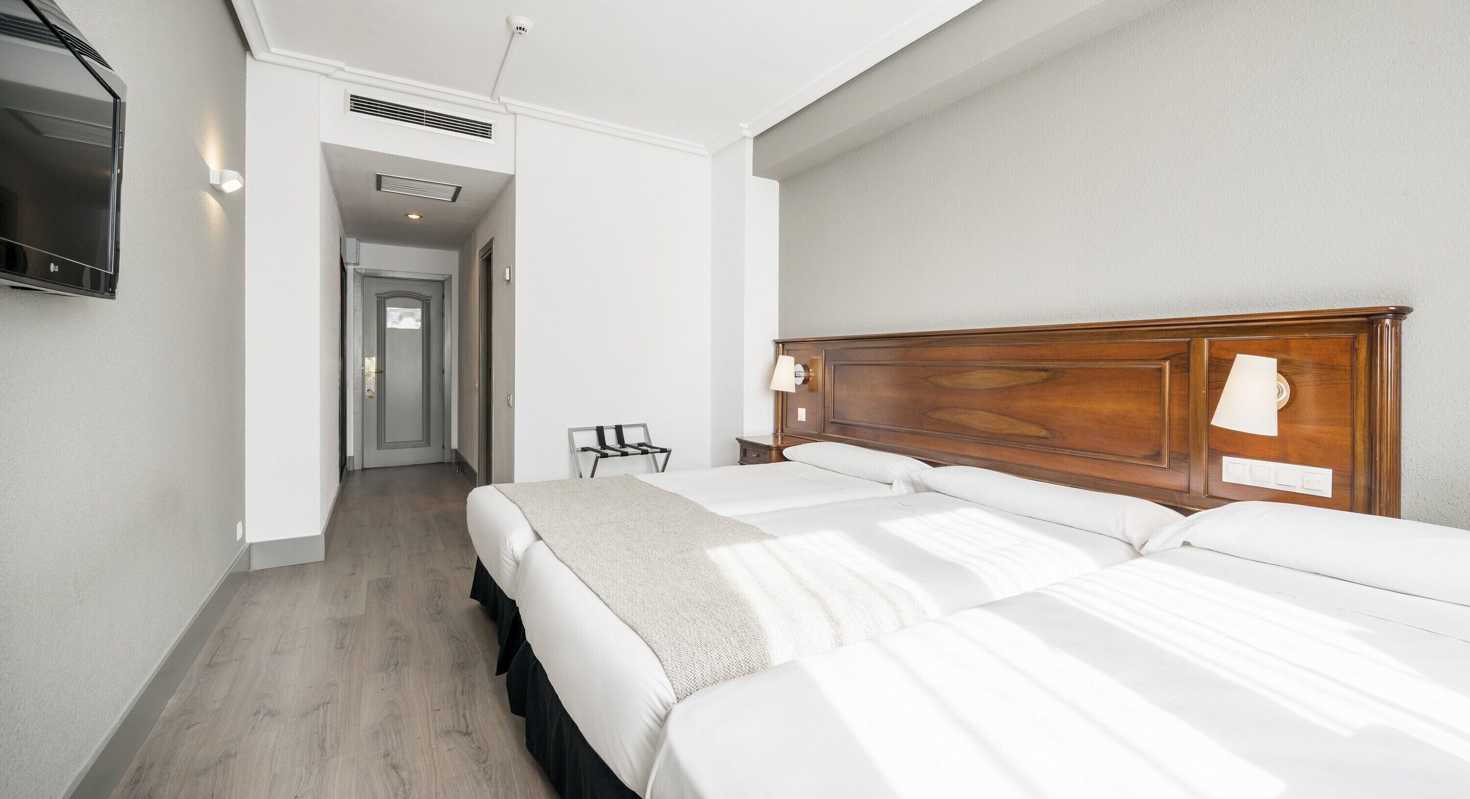 A hotel room with a wooden headboard, two beds, a flat-screen TV mounted on the wall, and a hallway leading to another room.