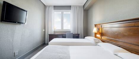 Una habitación de hotel con dos camas, un televisor de pantalla plana montado en la pared, un escritorio con una silla y una ventana con cortinas.