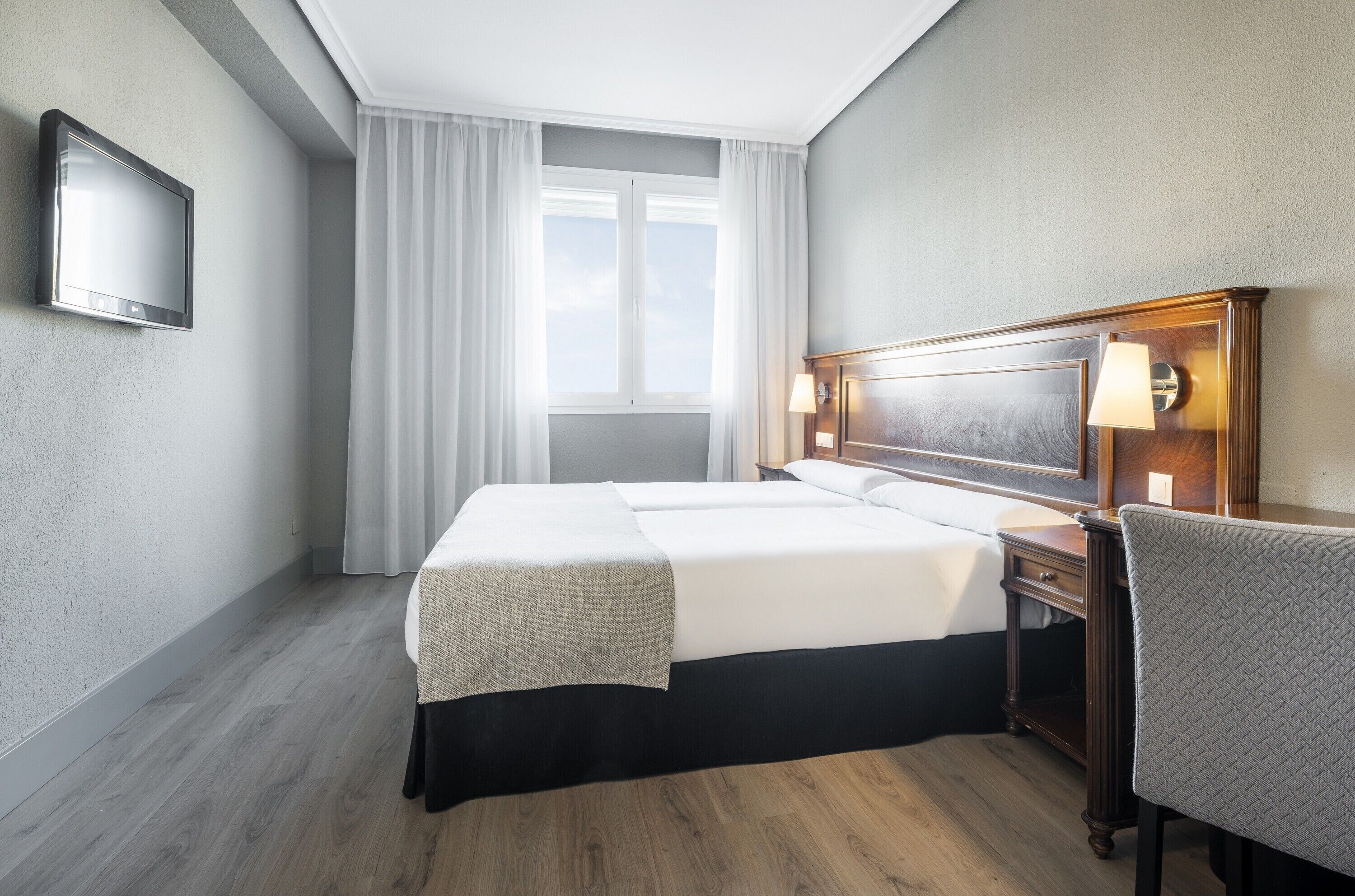 A hotel room with a bed, a desk with a lamp, a chair, a TV on the wall, and a window with curtains.