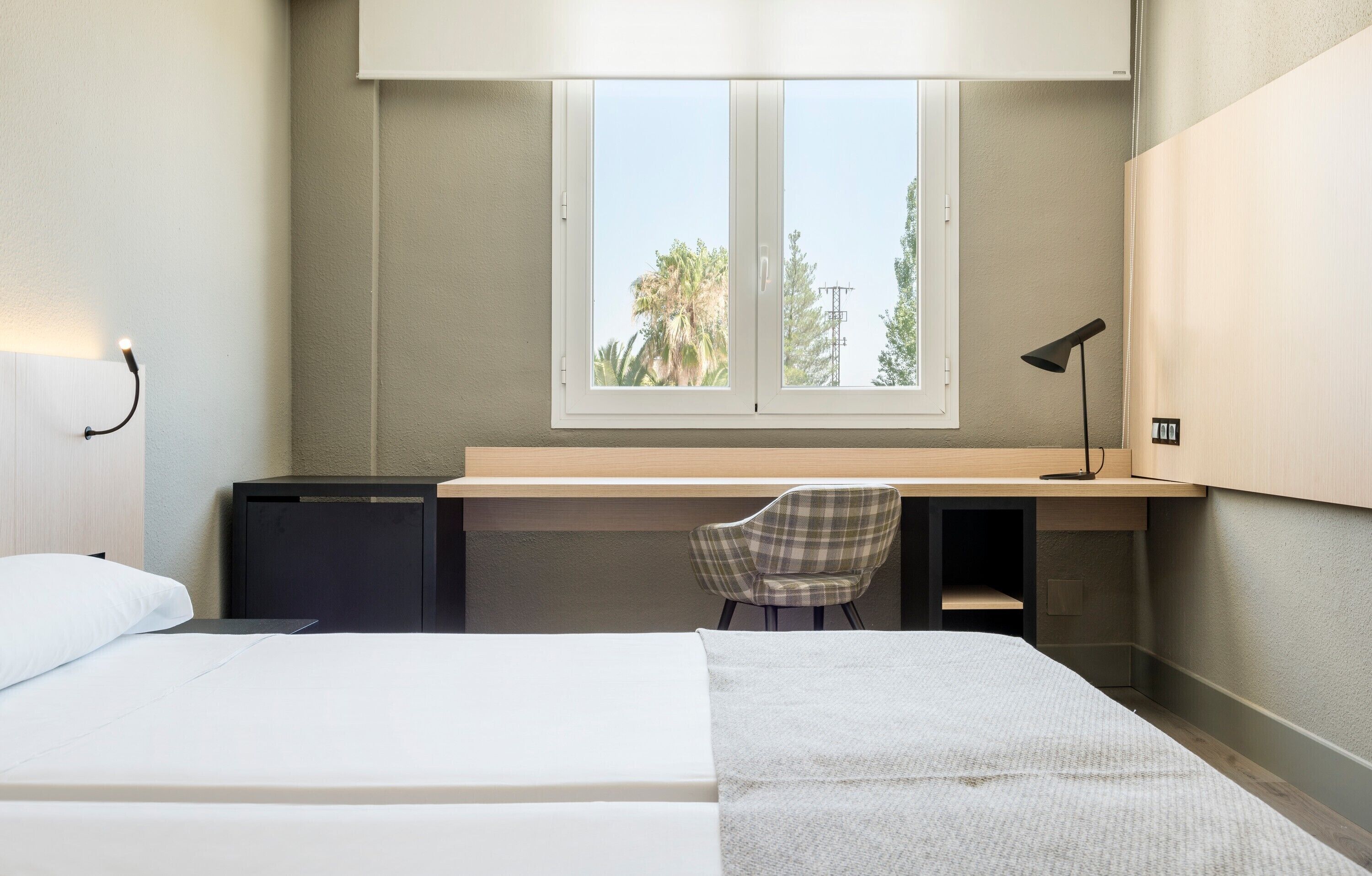 Un dormitorio con cama, escritorio, silla, ventana y lámpara.