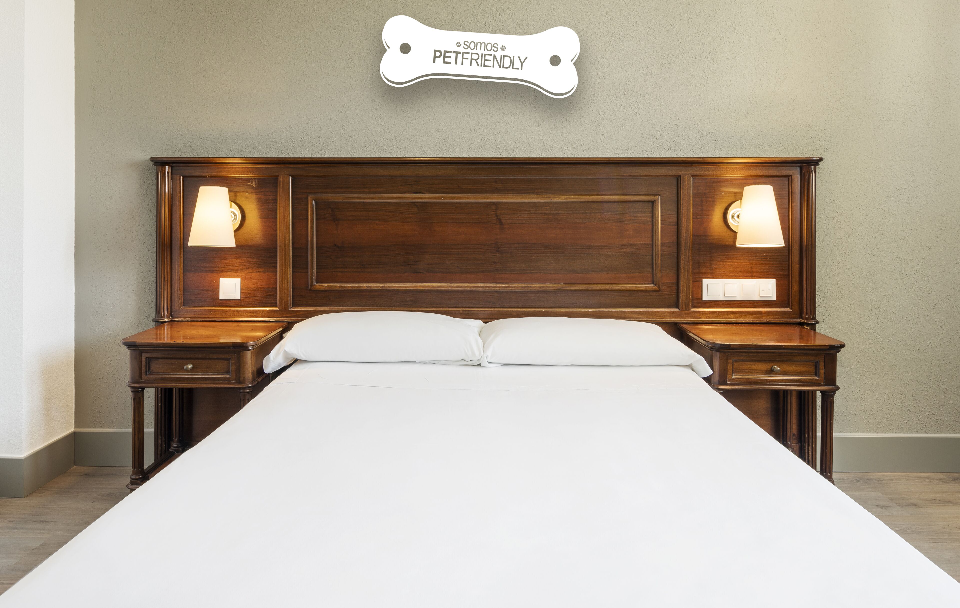 Una cama bien hecha con cabecera de madera, dos mesitas de noche con lámparas y un cartel sobre la cabecera que dice "Somos PetFriendly".