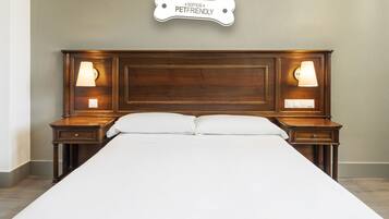 Una cama bien hecha con cabecera de madera, dos mesitas de noche con lámparas y un cartel sobre la cabecera que dice "Somos PetFriendly".