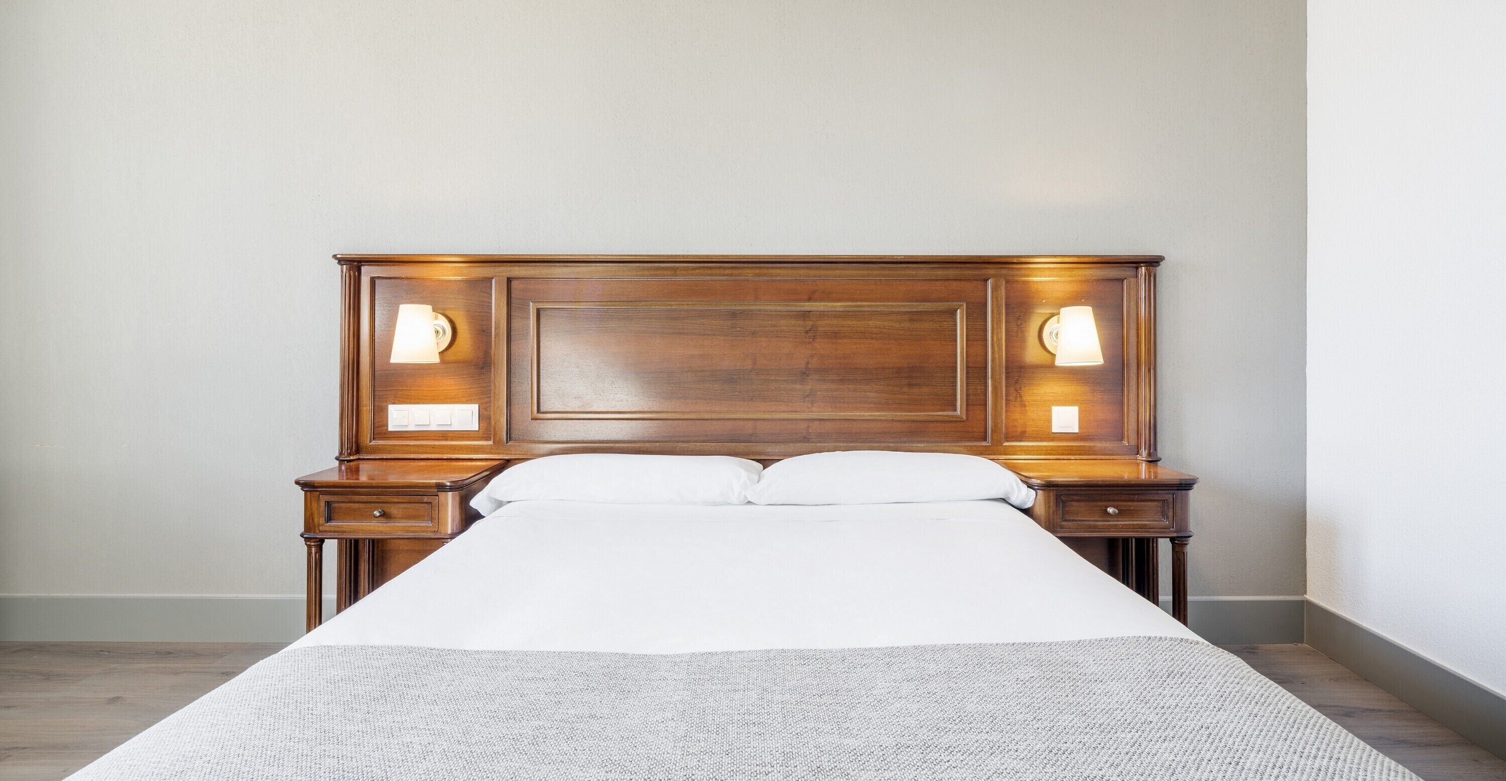 A neatly made bed with a wooden headboard, two bedside tables with lamps, and a plain wall.