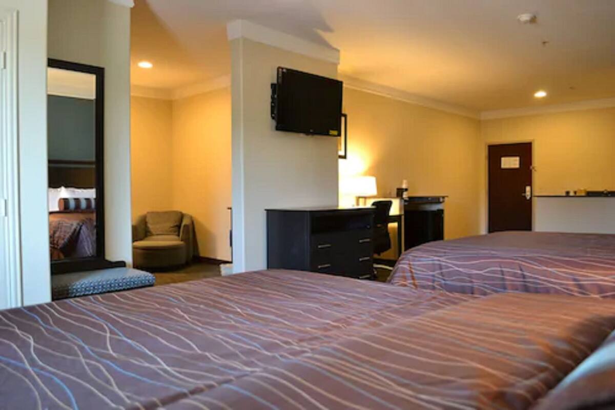 A hotel room with two beds, a TV mounted on the wall, a chair, a desk with a lamp, and a door.