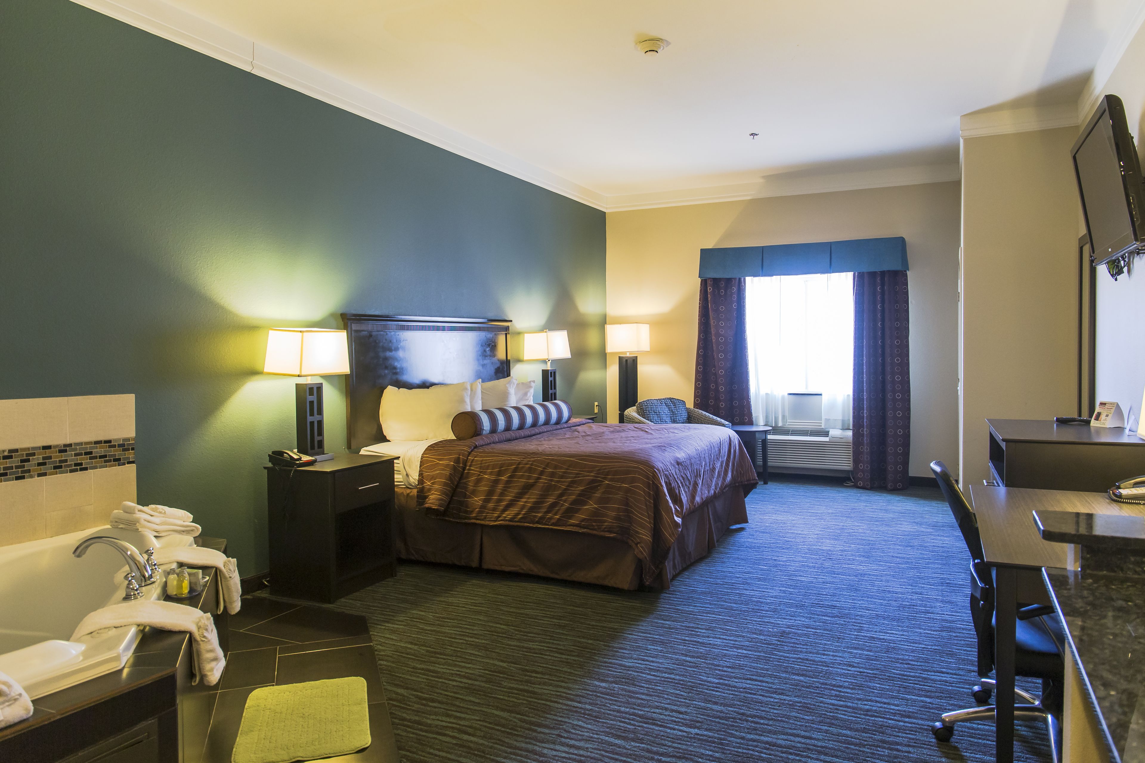 Une chambre d’hôtel comprenant un lit, un bureau, une chaise, une lampe de bureau, un téléphone, une salle de bain avec un lavabo et un miroir, et une fenêtre avec des rideaux.