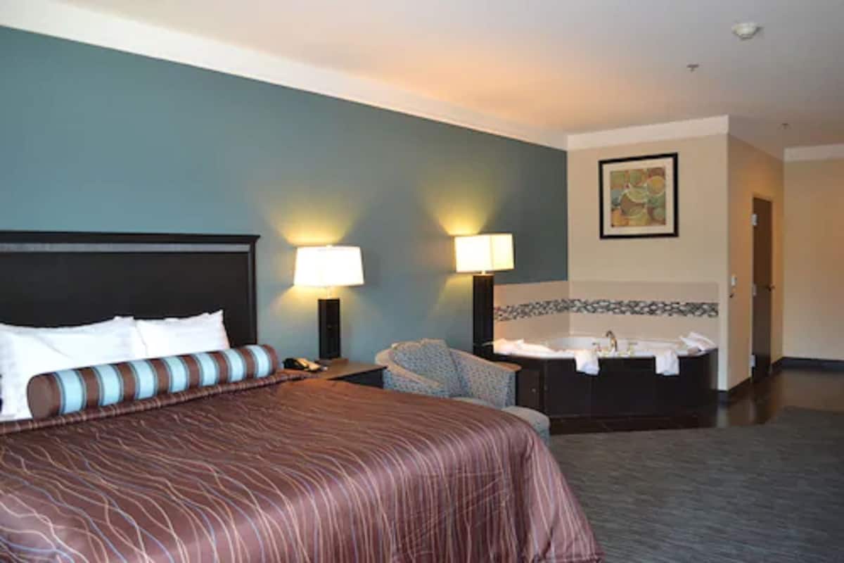 A hotel room with a large bed, two bedside lamps, a chair, a bathroom with a bathtub, and a painting on the wall.