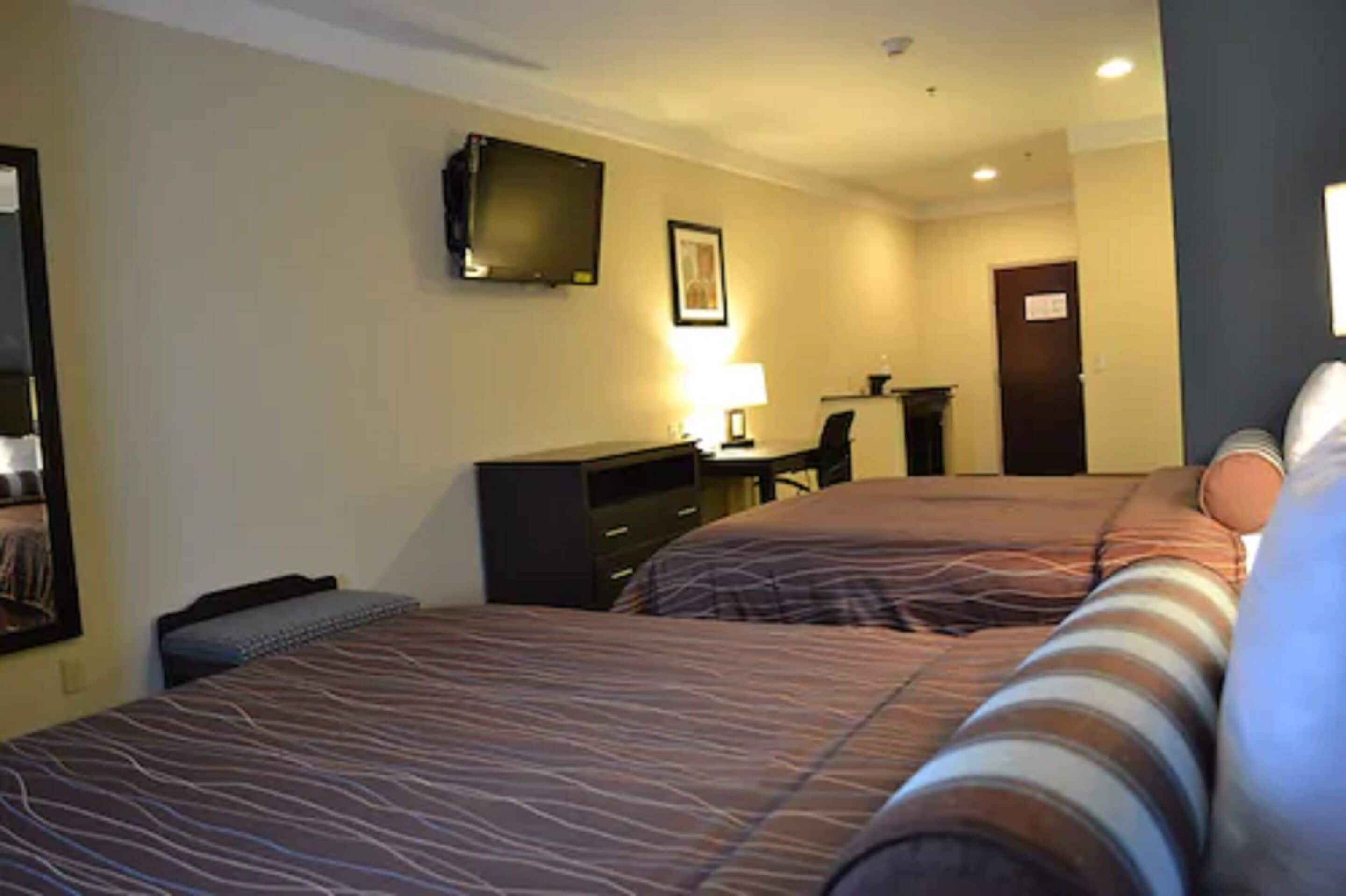 A hotel room with two beds, a desk, a chair, a television, and a lamp.