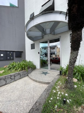 A modern building entrance with a curved glass door, a stone planter with plants, and a paved walkway. - Estrela da Água Fria (São Paulo)
