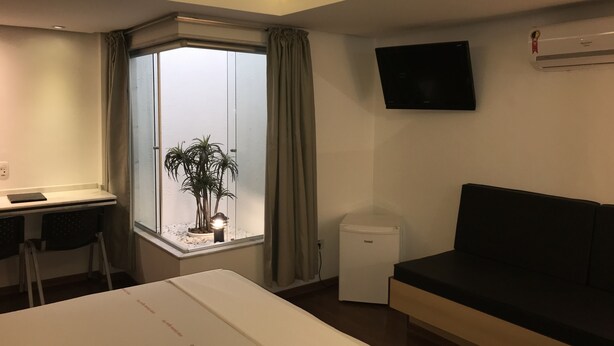 A modern hotel room with a bed, a desk, a chair, a small potted plant, a window with curtains, a wall-mounted TV, and a compact air conditioner. - Estrela da Água Fria (São Paulo)